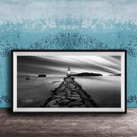 A black and white photograph of a lighthouse on a rocky pier, with a calm body of water and a cloudy sky in the background.