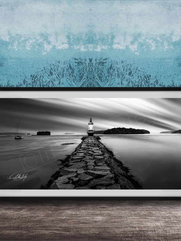 A black and white photograph of a lighthouse on a rocky pier, with a calm body of water and a cloudy sky in the background.