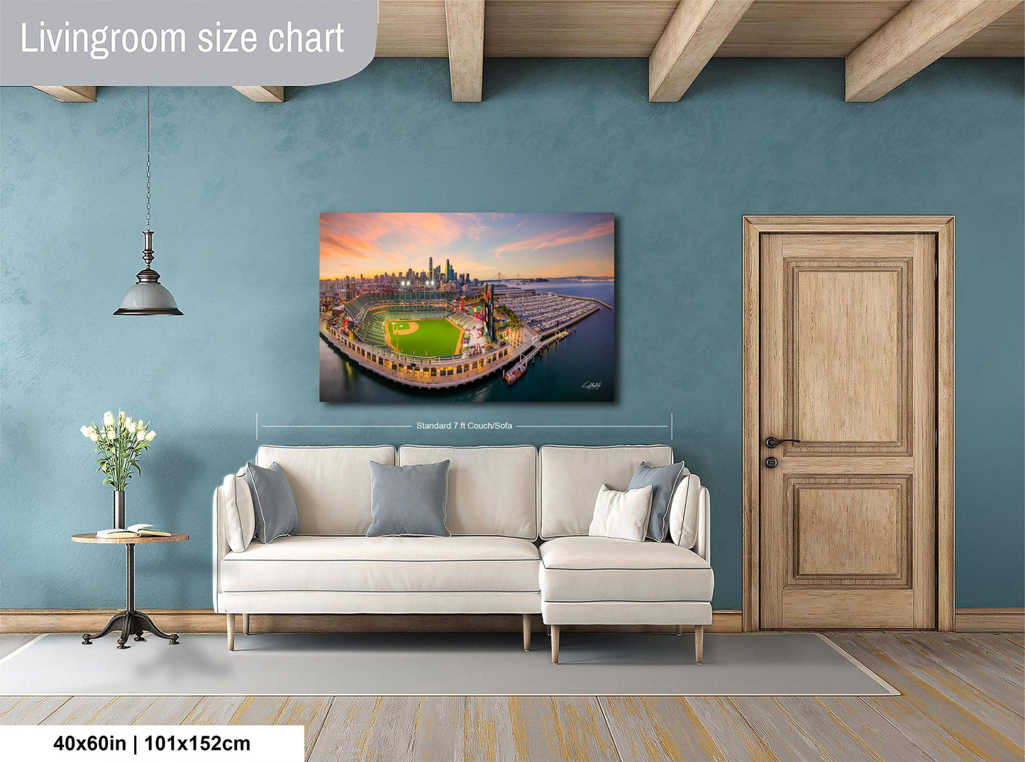 a living room with a large wall-mounted painting depicting a cityscape at sunset. The painting is framed and hangs above a white sofa. The room has a wooden door, a small table with a vase of flowers, and a wooden beam ceiling. The painting is approximately 40 inches wide and 115 centimeters tall.