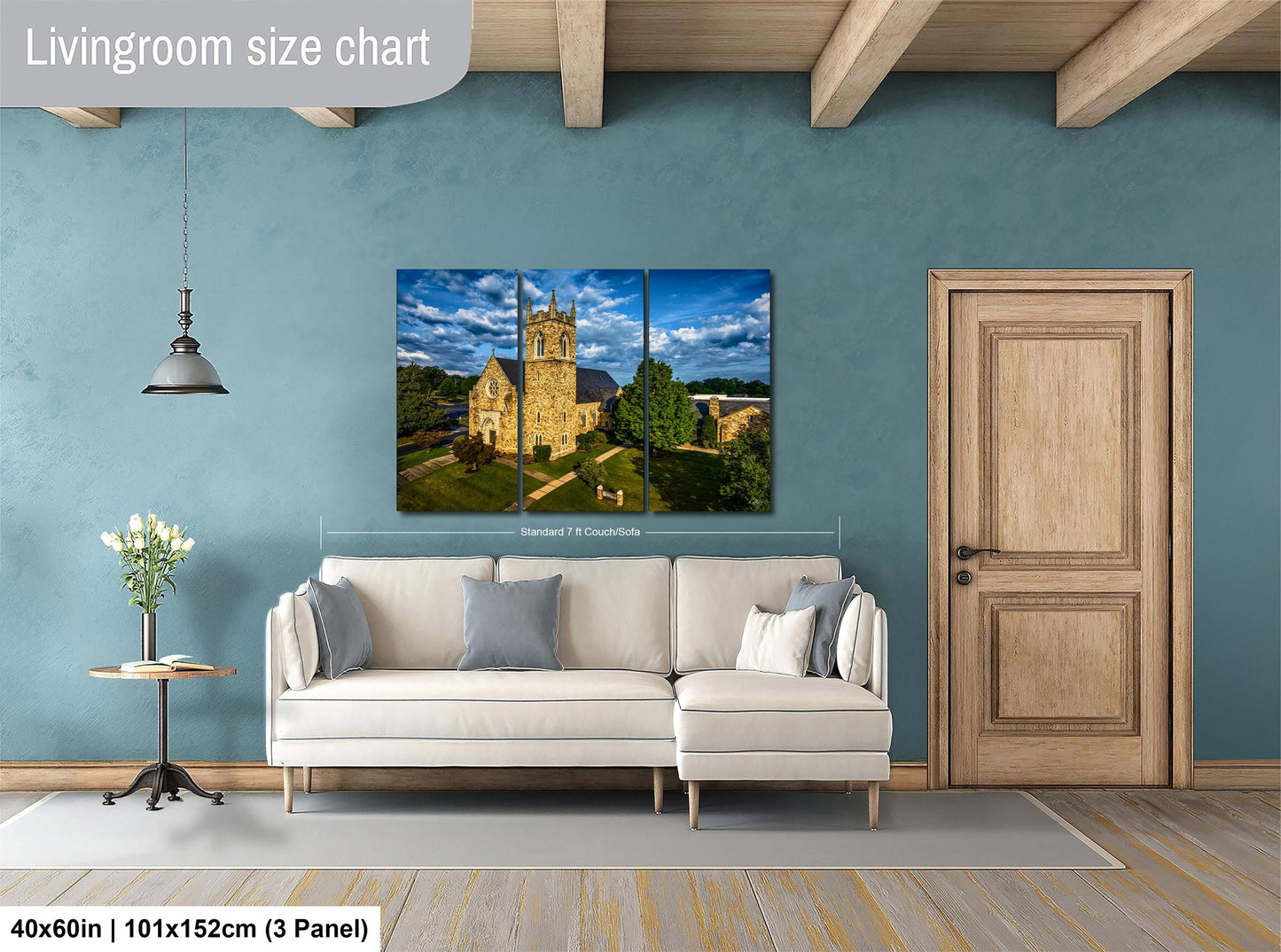 The image is a living room with a large painting of a castle on the wall. The painting is framed and hangs above a white sofa. The room has a wooden door and a small table with a vase of flowers. The wall is painted in a light blue color, and the floor is covered with a gray rug. The painting is approximately 40 inches wide and 101 inches tall, and it is mounted on a 3-panel panel.