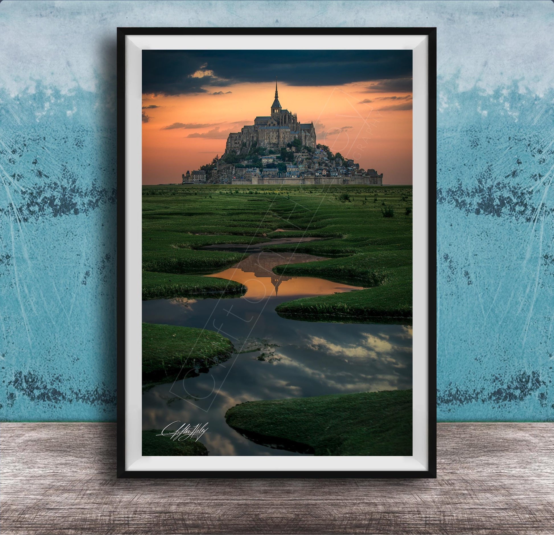 A framed photograph depicting the iconic Mont Saint-Michel, a medieval fortified island in Normandy, France, with its distinctive tower and spire against a vibrant sunset sky.