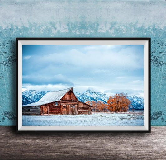 A large wooden barn with a sloping roof stands in a snowy field, with snow-capped mountains visible in the background.