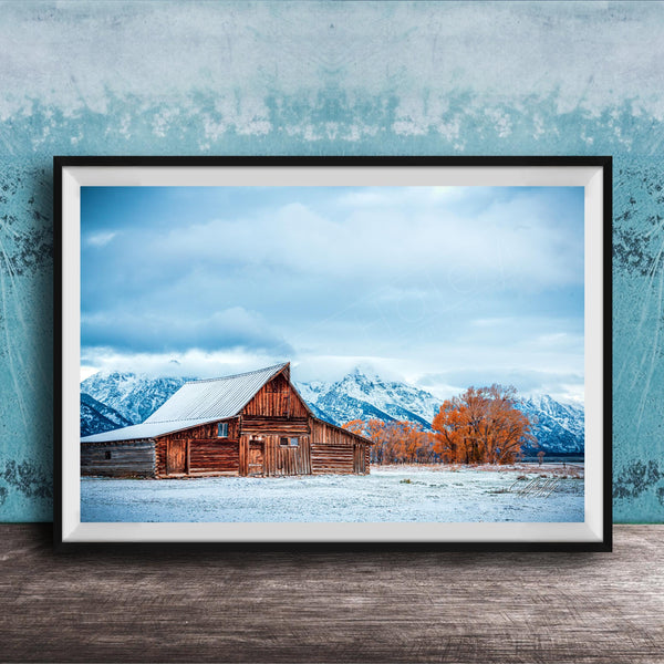 A large wooden barn with a sloping roof stands in a snowy field, with snow-capped mountains visible in the background.