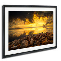 The image is a framed photograph depicting a rocky shoreline with a dramatic sunset in the background.