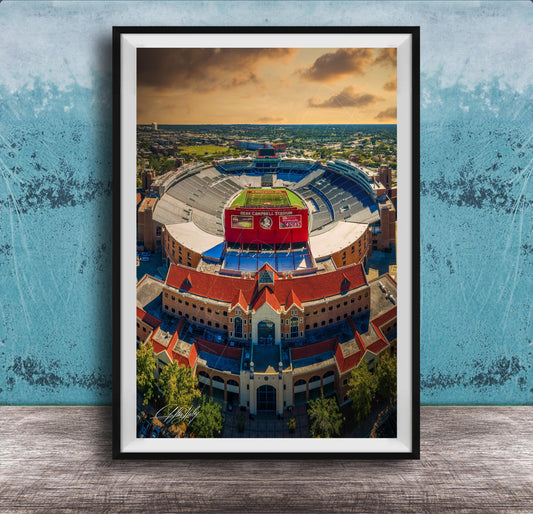 The image is an aerial view of a large stadium with a red roof, surrounded by buildings and a blue sky with clouds.