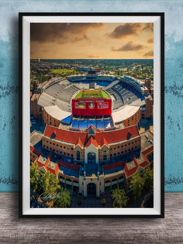 The image is an aerial view of a large stadium with a red roof, surrounded by buildings and a blue sky with clouds.