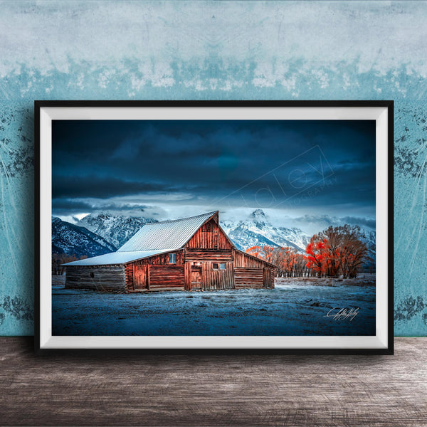 A rustic wooden barn stands in a snowy landscape, with a mountain range in the background under a dark, cloudy sky.