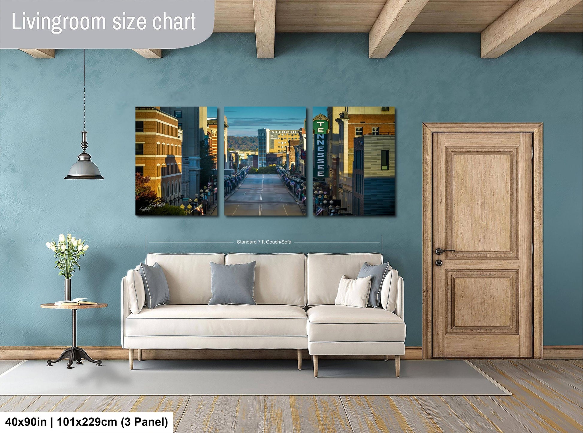 The image is a living room with a large white sofa and a wooden door. Above the sofa, there is a triptych of cityscape paintings depicting a city street with buildings and a bridge. The wall behind the sofa has a blue color, and there is a lamp hanging from the ceiling. The image is described as a living room size chart, indicating that it is a scale model or a representation of the actual living room size.