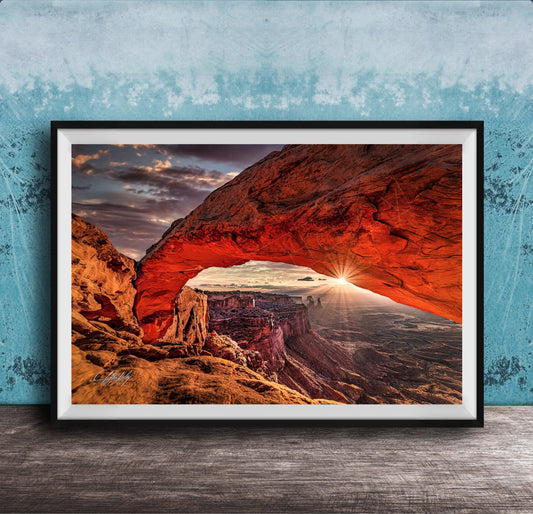 A framed photograph depicting a stunning natural landscape with a large arch formation in the distance, set against a backdrop of a cloudy sky and a sunset.