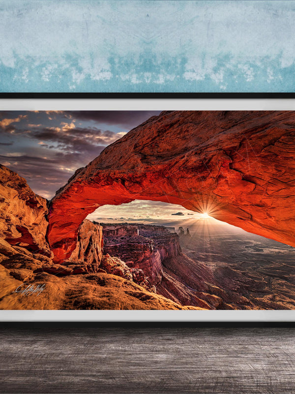 A framed photograph depicting a stunning natural landscape with a large arch formation in the distance, set against a backdrop of a cloudy sky and a sunset.