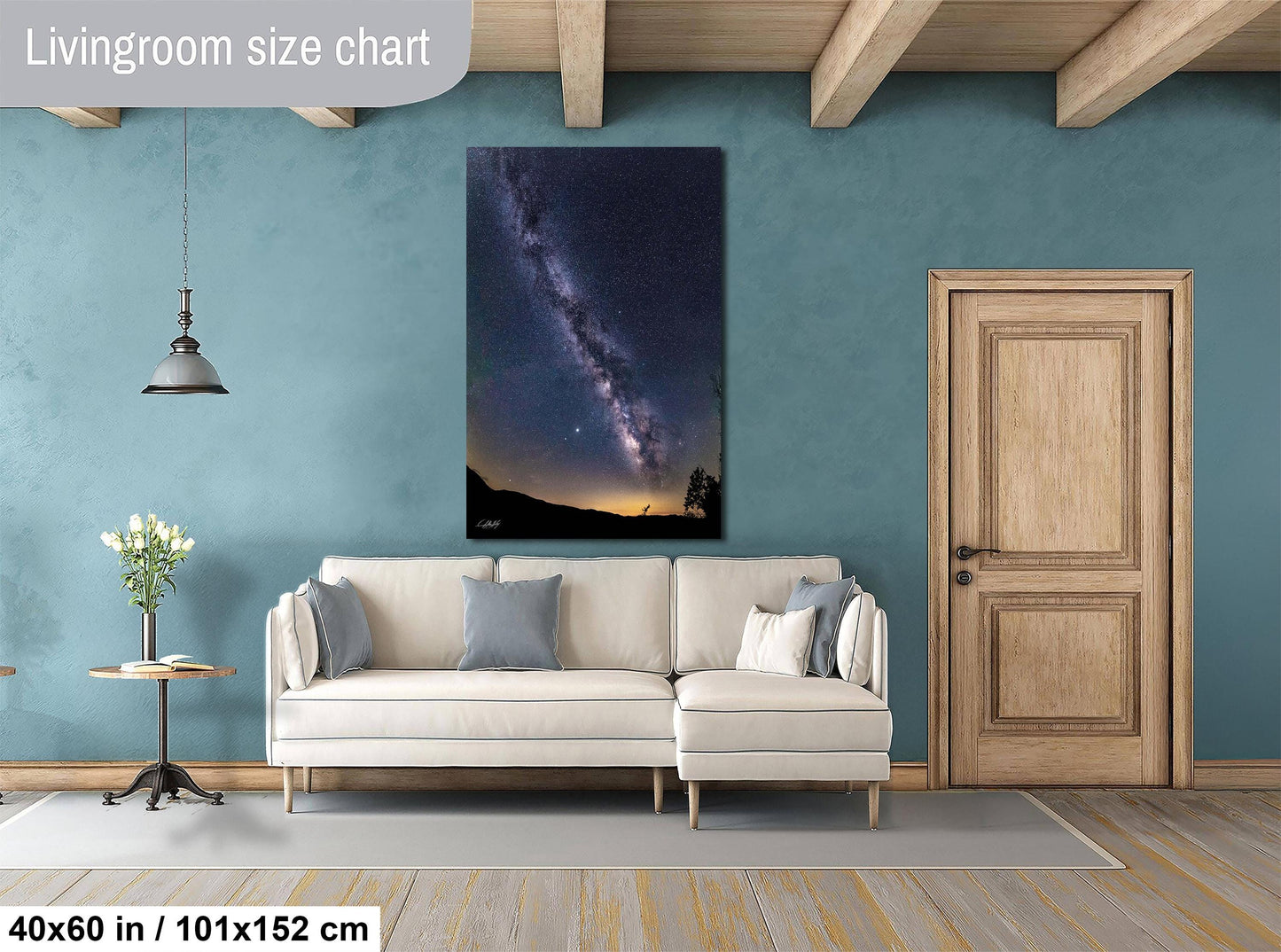 a living room with a large wall painting of a night sky featuring a galaxy and stars. The painting is hanging above a white sofa with two throw pillows. The room has a wooden door and a small table with a vase of flowers. The floor is covered with a gray rug.