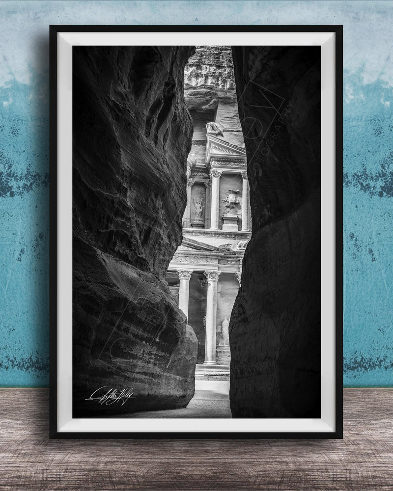 A black and white photograph of a narrow passage between two large rock formations, with a classical building visible through the opening.