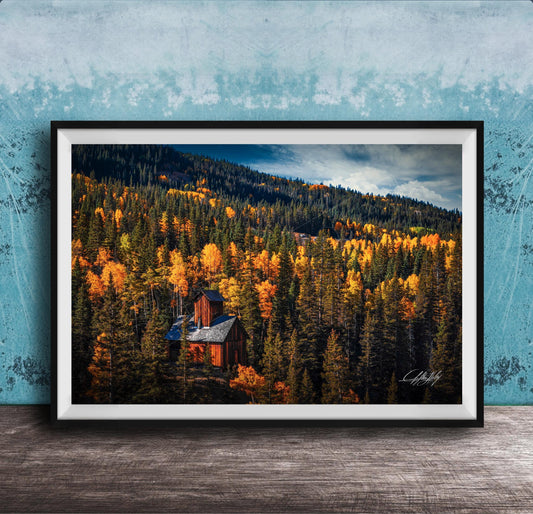 A framed photograph of a cabin nestled in a forest of trees with vibrant autumn foliage.