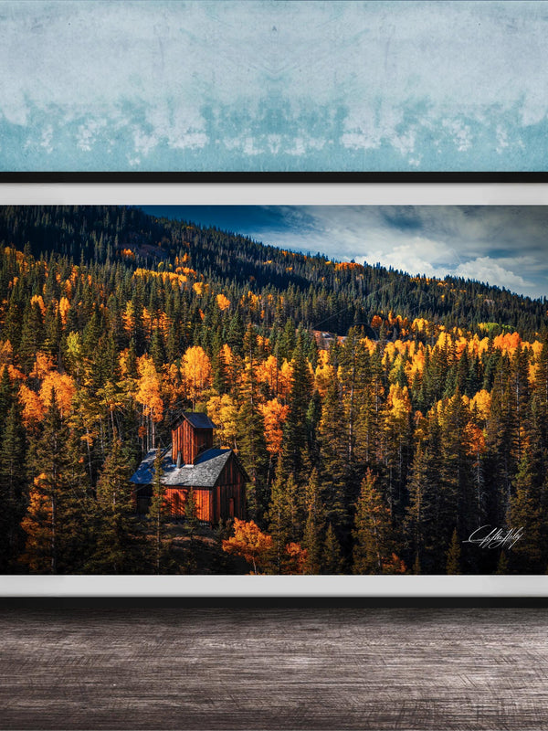 A framed photograph of a cabin nestled in a forest of trees with vibrant autumn foliage.