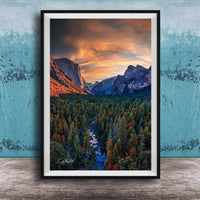 The image is a framed photograph of a mountainous landscape with a river running through it. The mountains are covered in trees, and the sky is filled with clouds, creating a dramatic and colorful scene.
