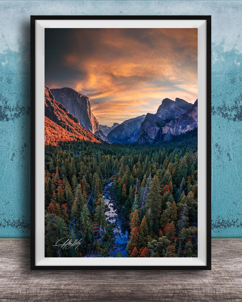 The image is a framed photograph of a mountainous landscape with a river running through it. The mountains are covered in trees, and the sky is filled with clouds, creating a dramatic and colorful scene.