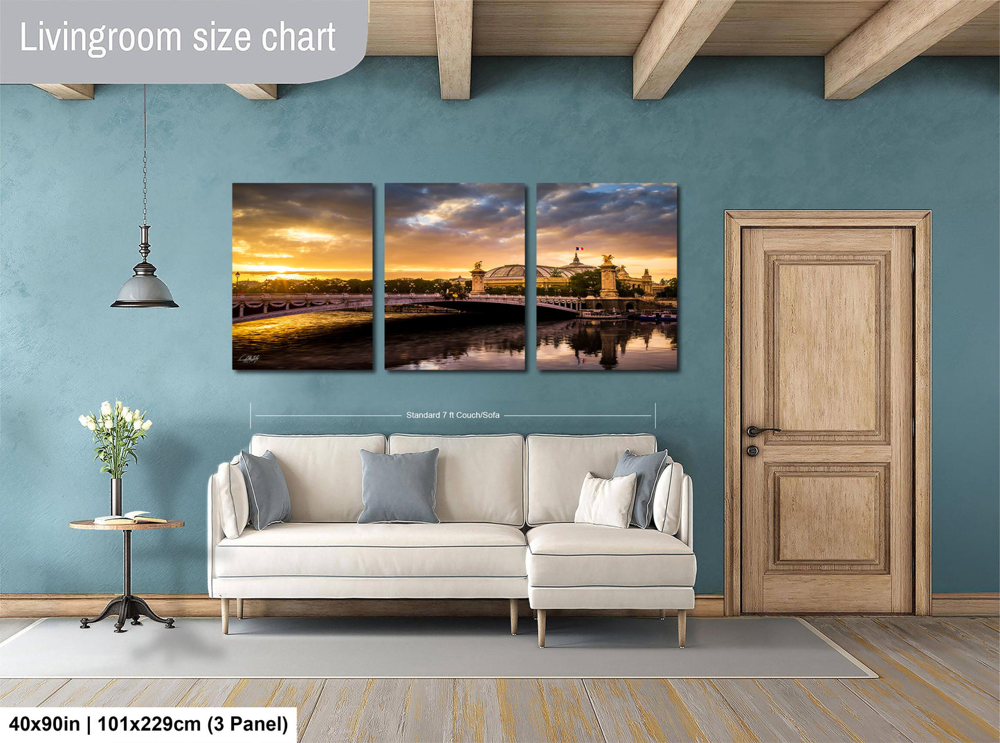 The image is a living room with a large wall art panel depicting a scenic view of a bridge and a sunset. The panel is divided into three sections, each showing a different part of the landscape. The living room has a white sofa and a wooden door, and the floor is covered with a gray rug.