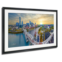The image is a framed aerial view of a city skyline, featuring a bridge, a river, and a sunset in the background.