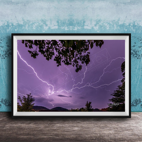 A framed photograph depicting a dramatic lightning storm with vibrant purple and blue hues illuminating the night sky, framed against a blue wall.