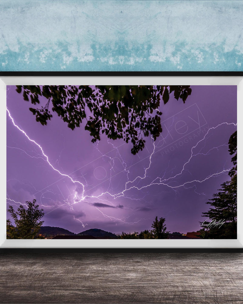 A framed photograph depicting a dramatic lightning storm with vibrant purple and blue hues illuminating the night sky, framed against a blue wall.
