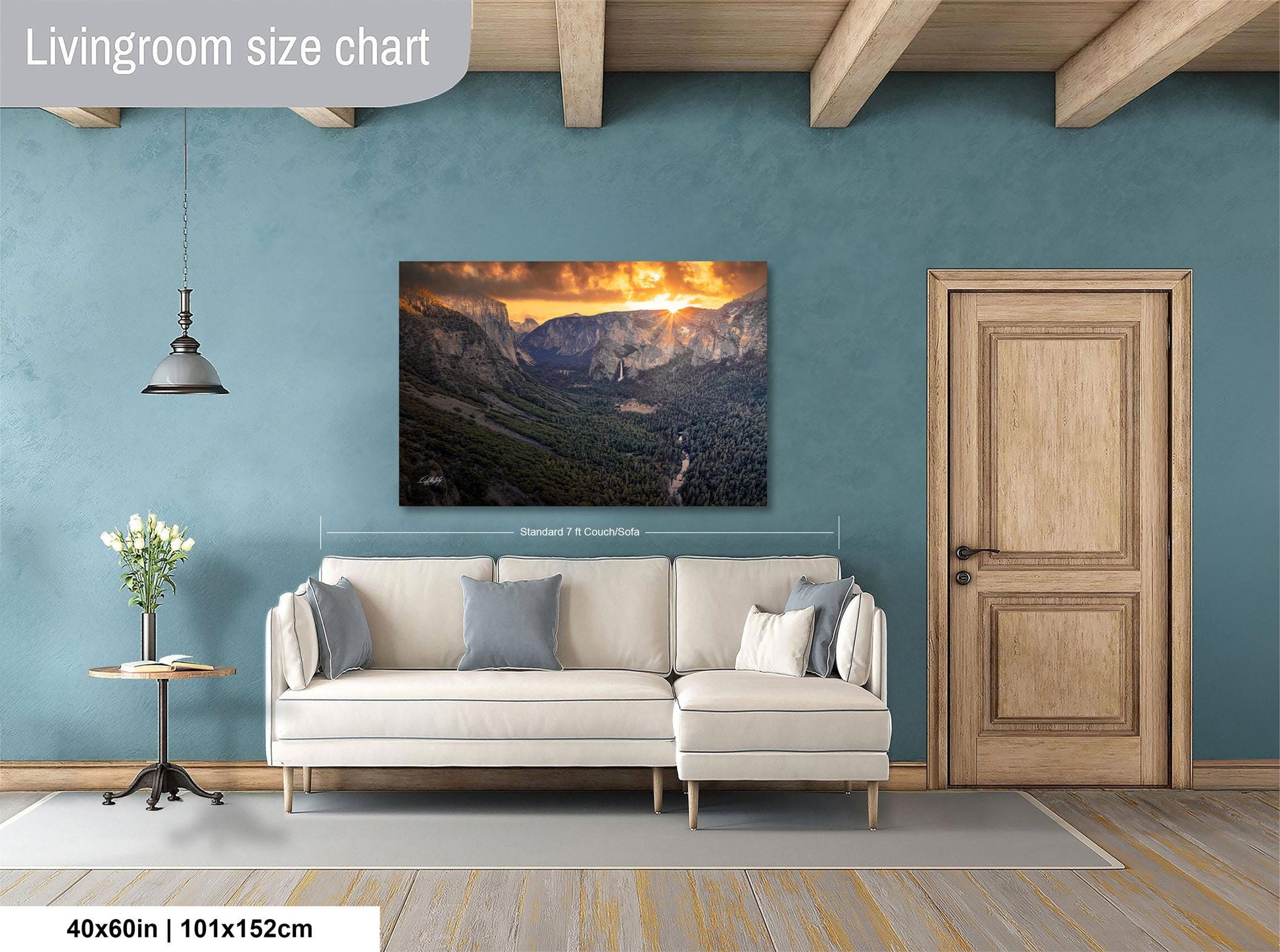 a living room with a large wall painting of a mountainous landscape, a white sofa, a wooden door, and a small table with a vase of flowers.