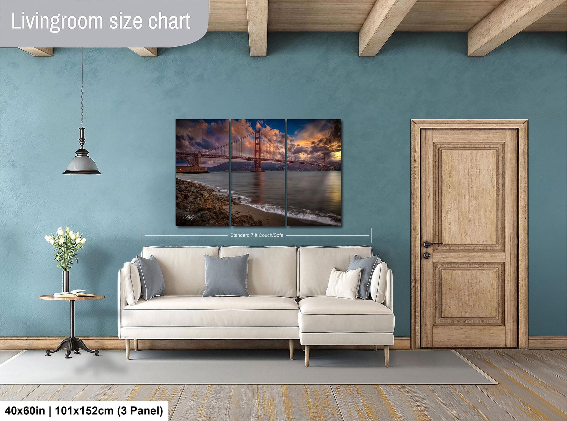 The image is a living room with a large, three-panel, landscape painting of the Golden Gate Bridge hanging on the wall. The painting is framed in a white frame and measures 40 inches by 101 inches by 150 centimeters. The room has a white sofa, a wooden door, and a small table with a vase of flowers.