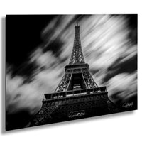 The image is a black and white photograph of the Eiffel Tower, a famous landmark in Paris, France. The tower is captured from a low angle, emphasizing its height and grandeur. The sky in the background is filled with clouds, creating a dramatic and moody atmosphere. The image is a collage of multiple images, and the artist's signature is visible in the bottom right corner.