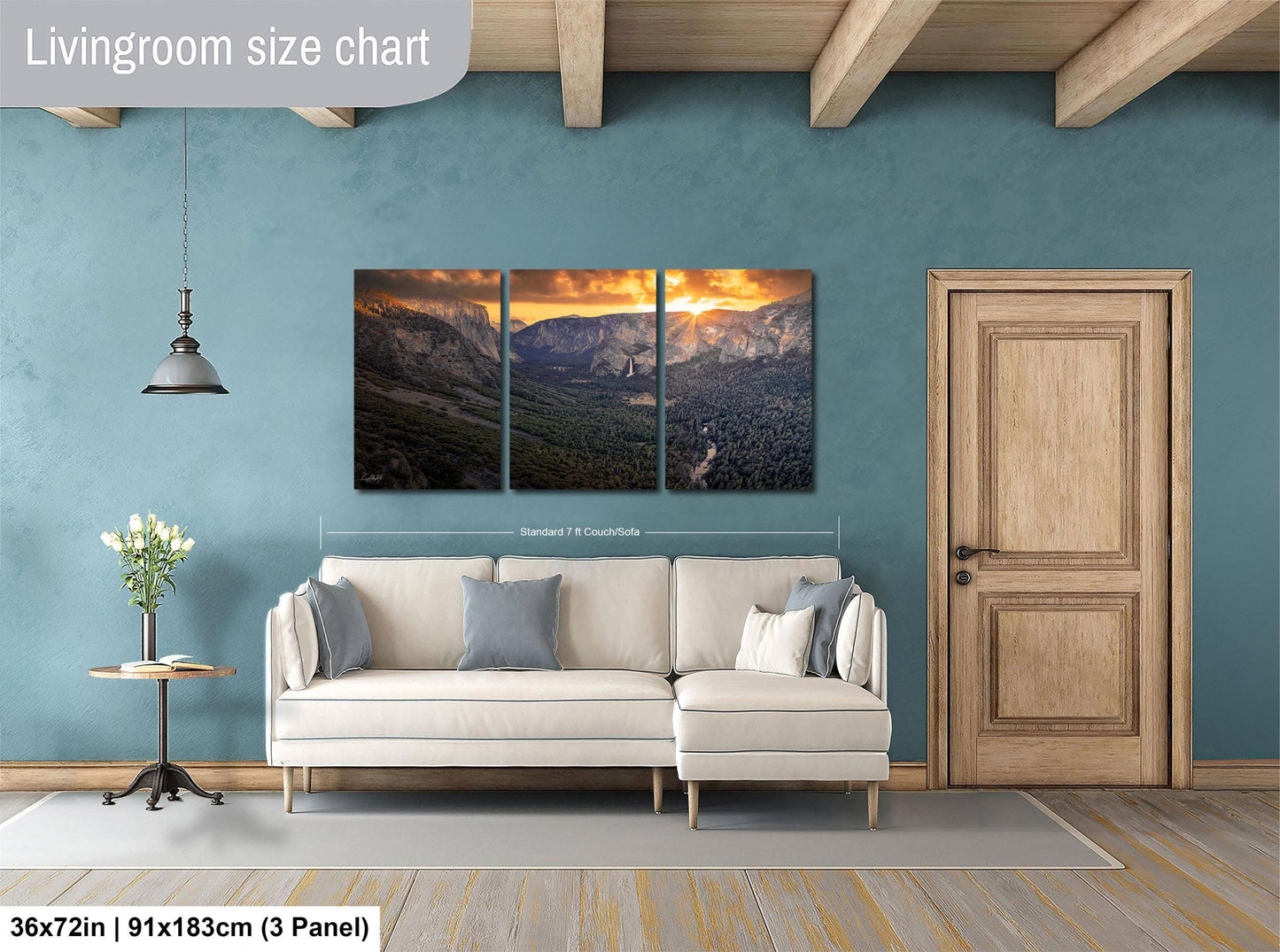 The image is a living room with a large, three-panel landscape painting hanging on the wall. The painting depicts a mountainous scene with a sunset in the background. The room has a white sofa and a wooden door, and there is a vase of flowers on a table. The painting is approximately 36 inches wide and 15 inches tall, and it is mounted on a wooden beam.