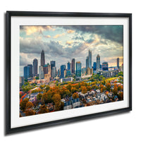 a cityscape with tall buildings and skyscrapers, surrounded by trees with autumn foliage. The sky is cloudy, creating a dramatic backdrop for the city skyline.