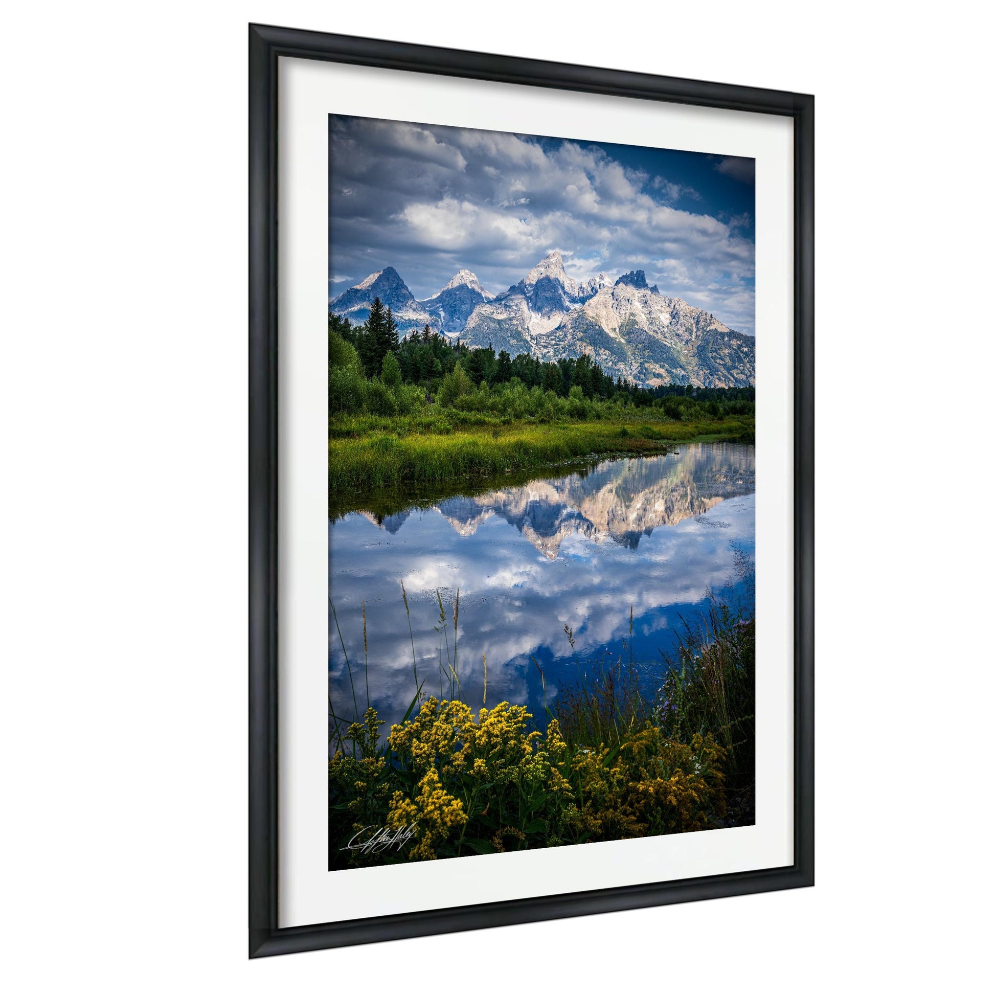a serene landscape with a calm lake reflecting the majestic snow-capped mountains in the background. The mountains are surrounded by lush green forests, creating a picturesque scene. The sky above is a clear blue, dotted with fluffy white clouds. The image is framed and appears to be a high-quality, professional-looking photograph.