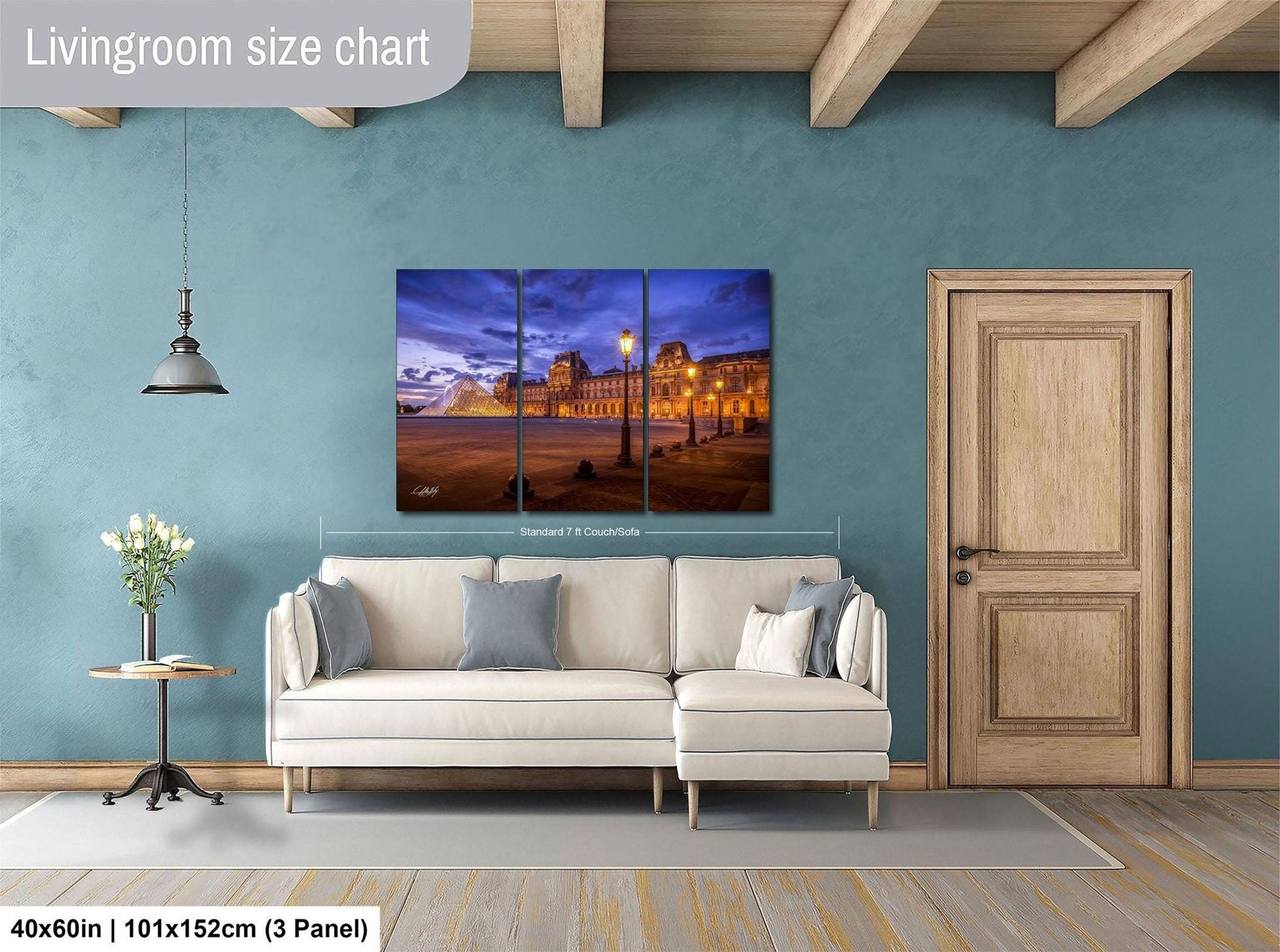 The image is a living room with a large, high-quality, three-panel print of a cityscape at night. The print is mounted on the wall above a white sofa, and there is a wooden door and a small table with a vase of flowers nearby. The print is approximately 40 inches wide and 101.5 inches tall, with a 3-panel layout.