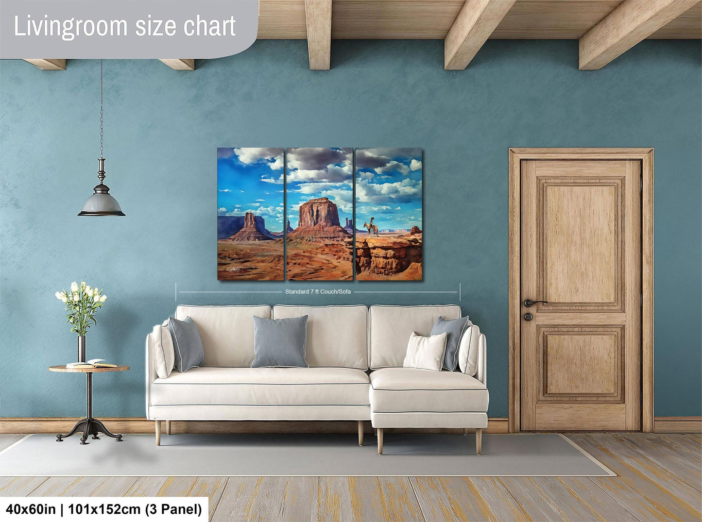 The image is a livingroom with a large painting of a desert landscape hanging on the wall. The painting is divided into three panels, depicting a desert scene with mountains and a clear blue sky. The livingroom has a white sofa and a wooden door, and there is a vase of flowers on a side table. The painting is approximately 40 inches wide and 101 inches tall, and it is mounted on a wooden panel.