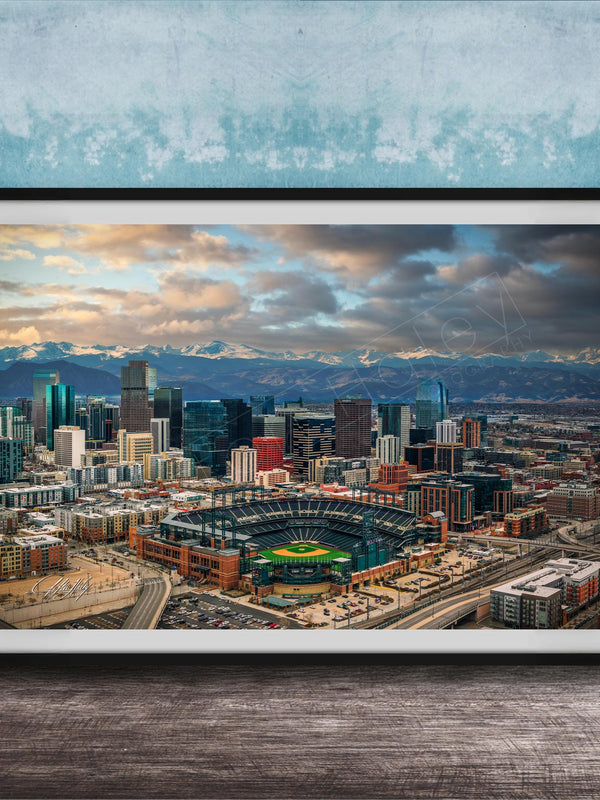 The image is a framed aerial view of a city skyline, featuring a baseball stadium and mountains in the background.
