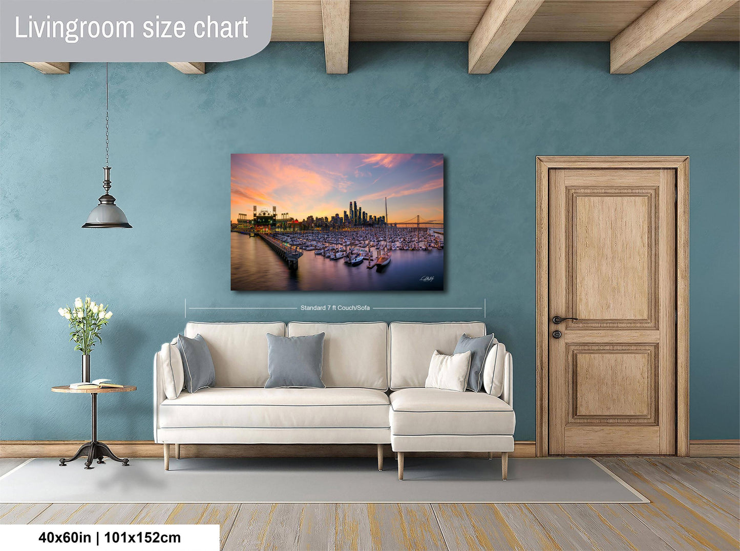 a living room with a large wall-mounted painting of a city skyline at sunset. The painting is framed and hangs above a white sofa, which is positioned in front of a wooden door. The room has a wooden floor and a small table with a vase of flowers. The wall is painted in a light blue color, and the overall layout and design of the room create a cozy and inviting atmosphere.