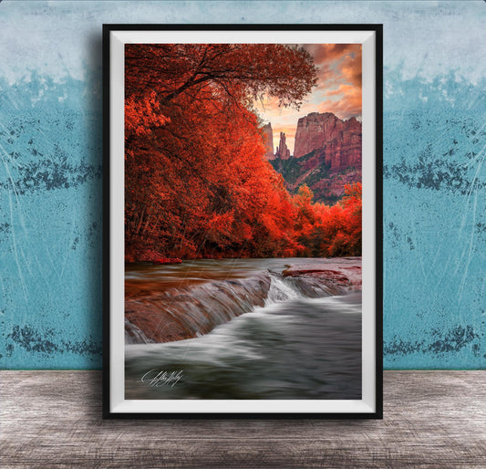 A framed photograph depicting a serene landscape with a waterfall cascading over rocks, surrounded by vibrant red foliage and set against a backdrop of towering red rock formations.