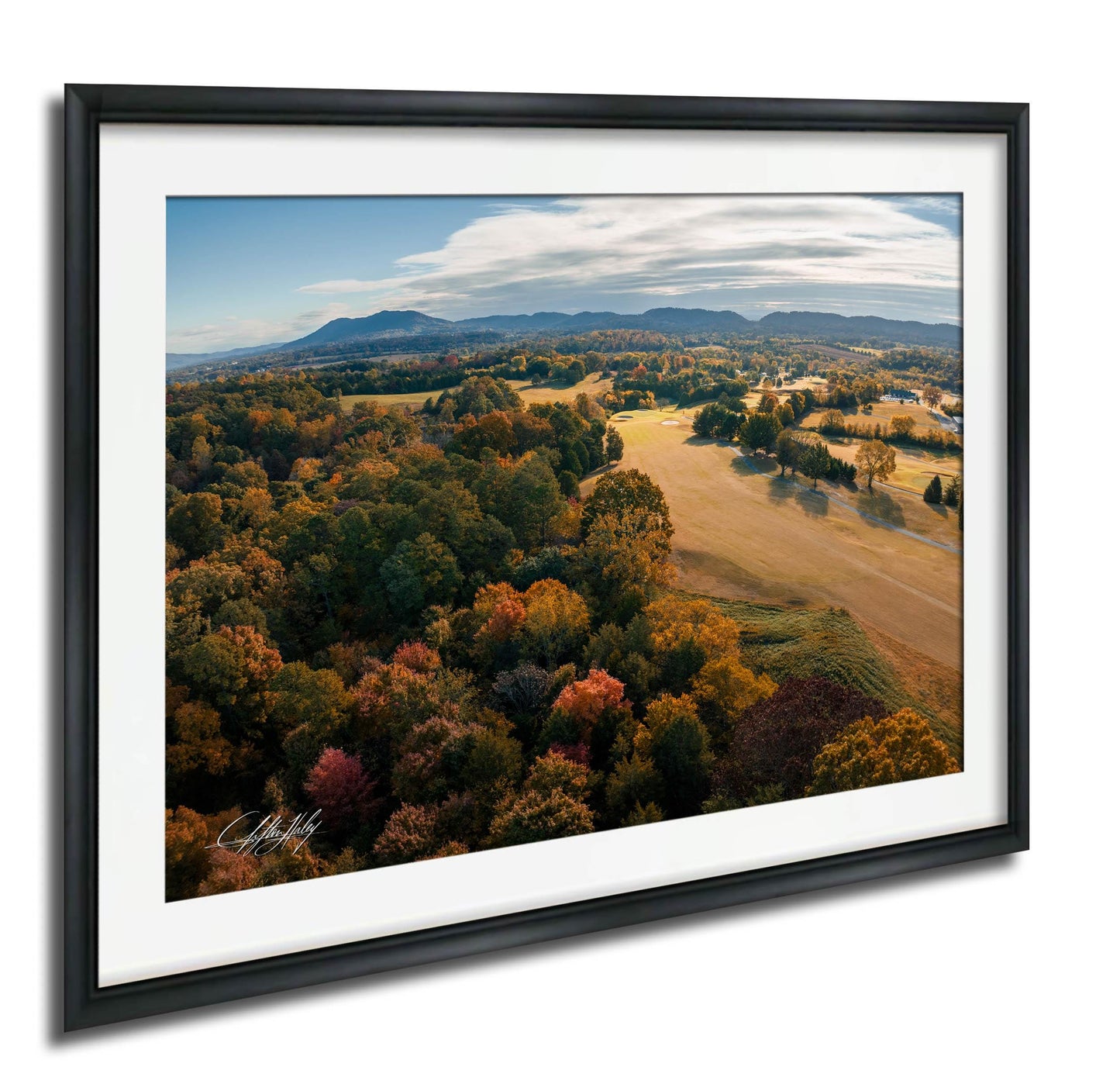 The image is a framed aerial view of a scenic landscape with a mix of trees in autumn colors, rolling hills, and a clear blue sky with clouds.