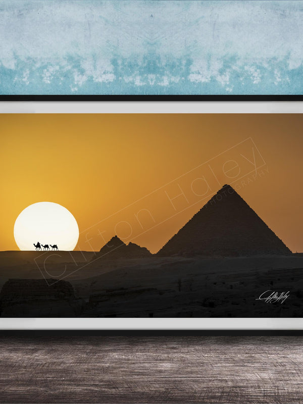 A framed photograph depicting the silhouette of the Great Pyramids of Giza against a vibrant sunset sky, with camels in the foreground.