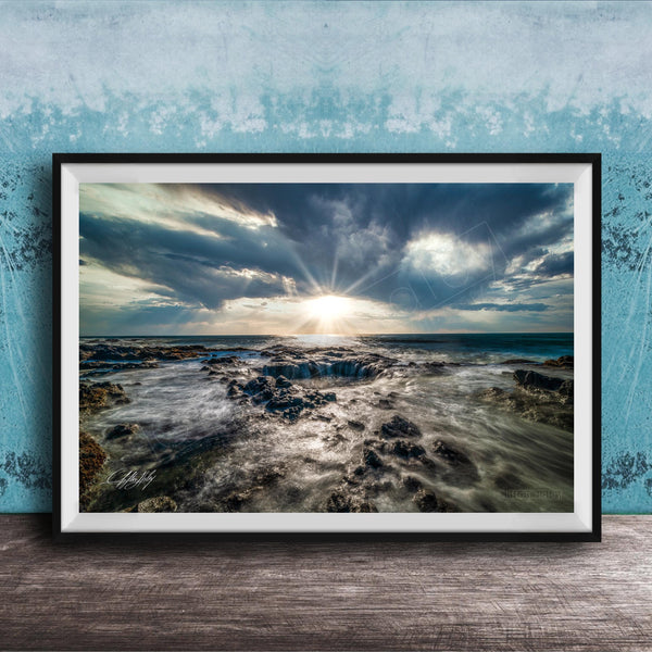The image is a framed photograph of a rocky coastline with crashing waves, set against a backdrop of a cloudy sky and the sun peeking through the clouds.