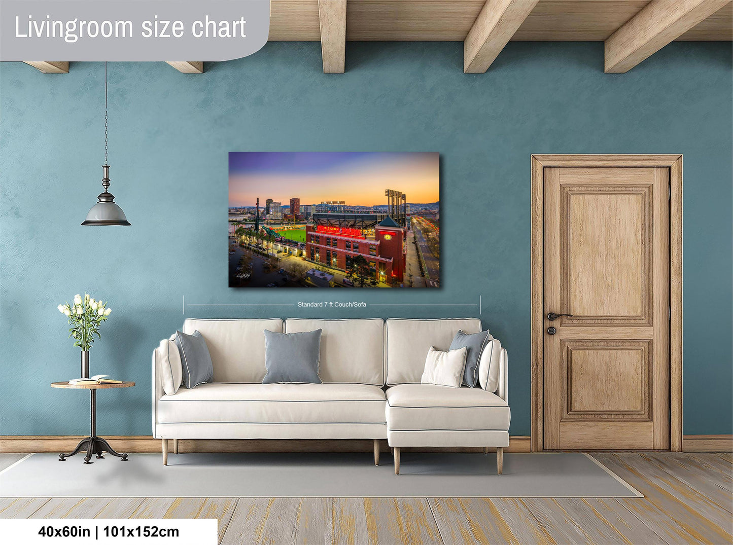 a living room with a large wall-mounted painting depicting a cityscape at sunset. The painting is framed and hangs above a white sofa. The room has a wooden door, a small table with a vase of flowers, and a wooden beam ceiling. The painting is approximately 40 inches wide and 115 centimeters tall.