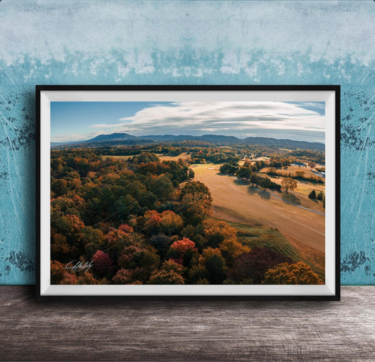 The image is a framed aerial view of a scenic landscape with a mix of trees, fields, and mountains in autumn colors.