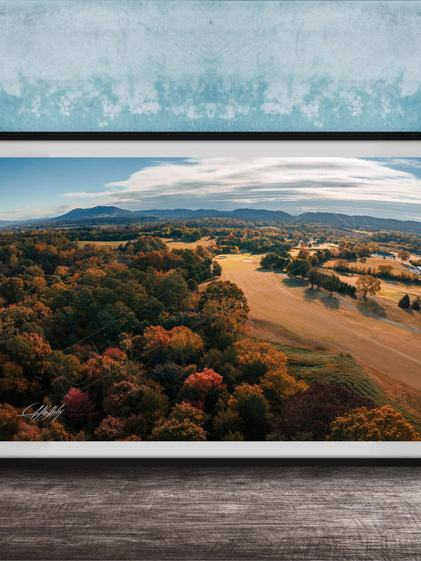 The image is a framed aerial view of a scenic landscape with a mix of trees, fields, and mountains in autumn colors.