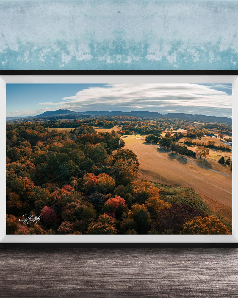 The image is a framed aerial view of a scenic landscape with a mix of trees, fields, and mountains in autumn colors.