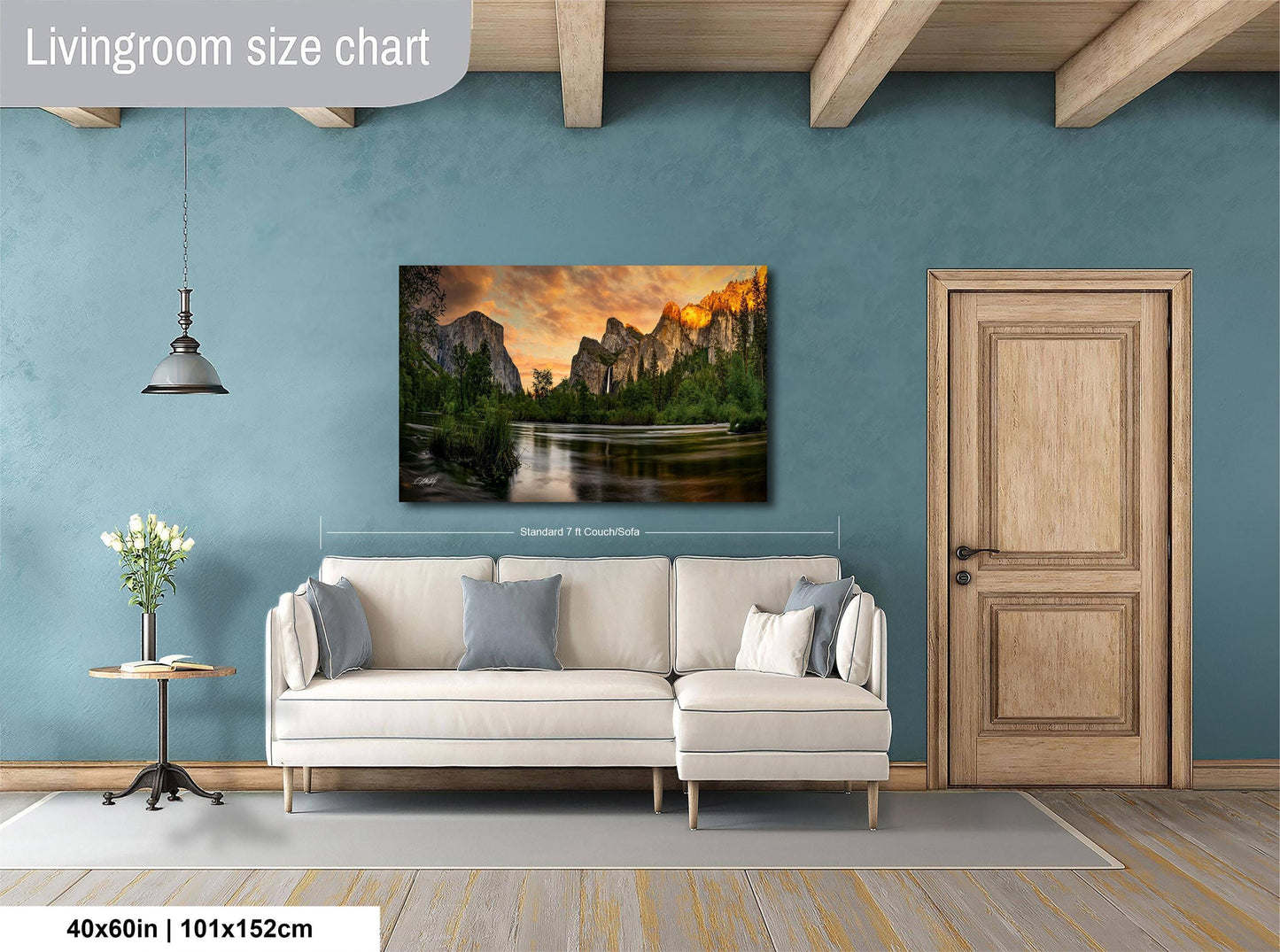 a living room with a large wall painting of a scenic landscape featuring mountains and a lake. The painting is hung above a white sofa, which is positioned in front of a wooden door. The room has a wooden floor and a small table with a vase of flowers. The wall painting is approximately 40 inches wide and 115 centimeters tall.