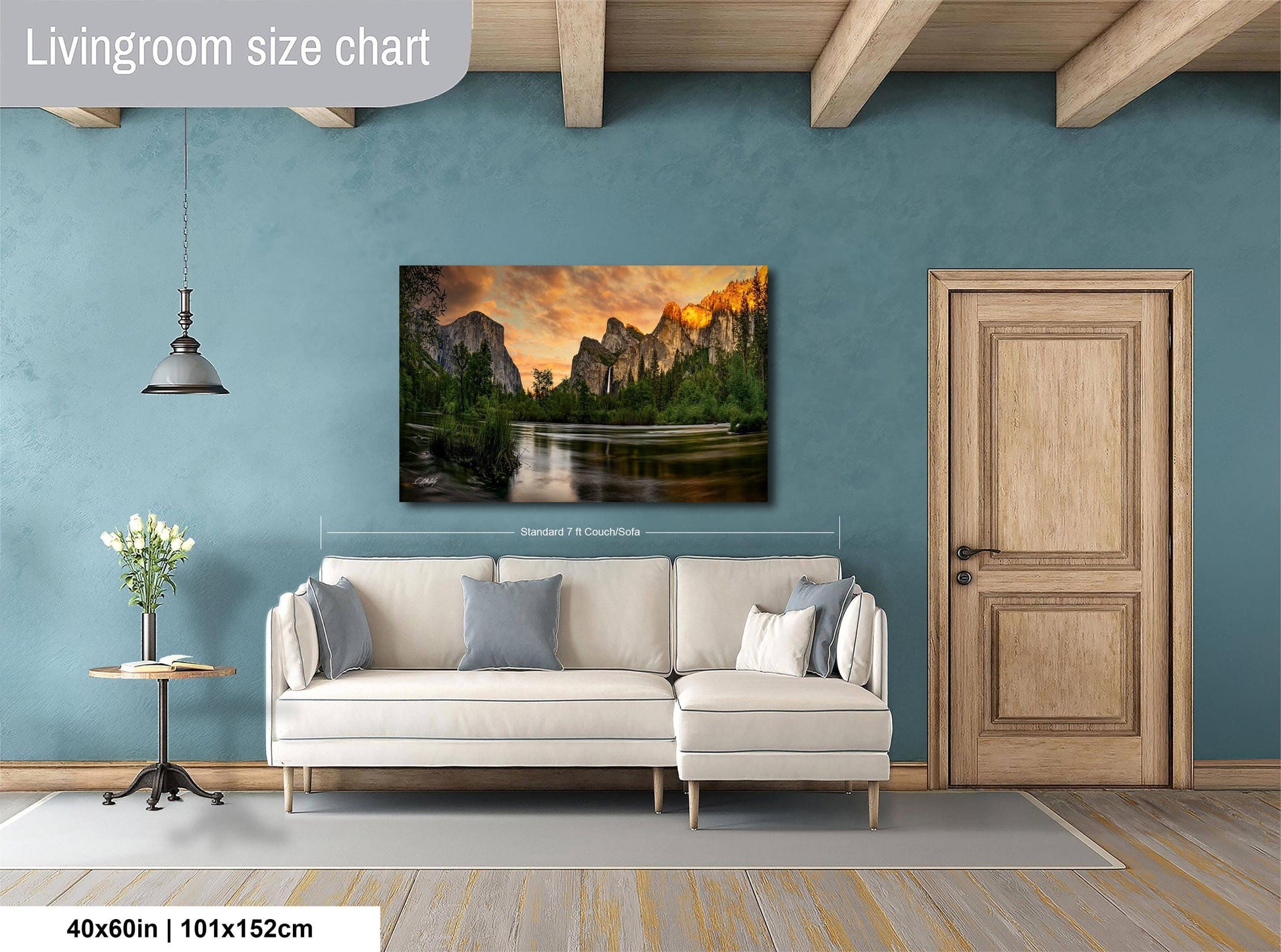 a living room with a large wall painting of a scenic landscape featuring mountains and a lake. The painting is hung above a white sofa, which is positioned in front of a wooden door. The room has a wooden floor and a small table with a vase of flowers. The wall painting is approximately 40 inches wide and 115 centimeters tall.