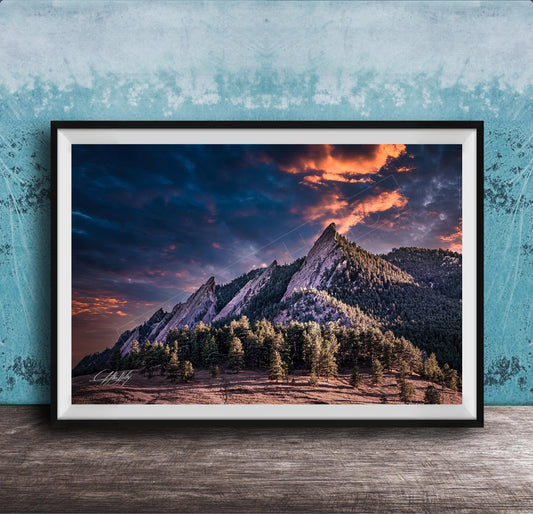 The image is a framed photograph of a mountainous landscape with a dramatic sky at sunset. The mountains are covered in trees and appear to be covered in snow, creating a serene and picturesque scene. The sky is filled with vibrant colors, including shades of orange, pink, and blue, indicating the sun setting over the mountains. The photograph is framed and displayed on a wooden surface, likely on a wall.