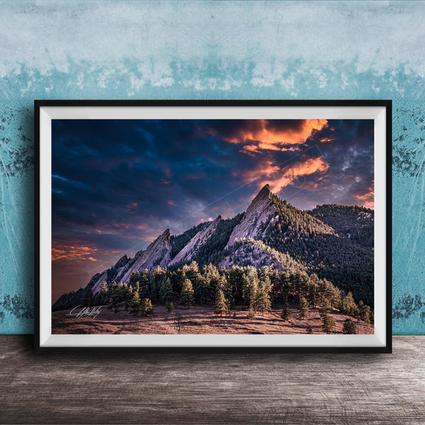 The image is a framed photograph of a mountainous landscape with a dramatic sky at sunset. The mountains are covered in trees and appear to be covered in snow, creating a serene and picturesque scene. The sky is filled with vibrant colors, including shades of orange, pink, and blue, indicating the sun setting over the mountains. The photograph is framed and displayed on a wooden surface, likely on a wall.