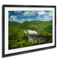A steam train crossing a stone bridge in a picturesque countryside setting with rolling hills and a blue sky with clouds.
