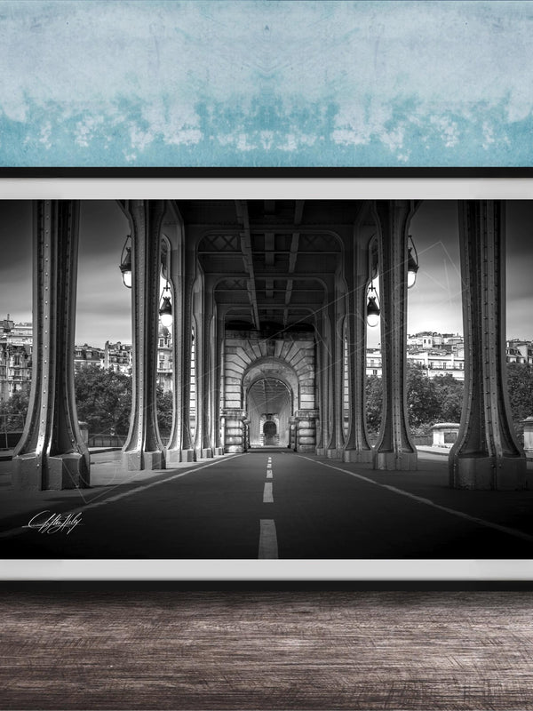 A black and white photograph of a long, narrow bridge with tall columns, leading to a central archway. The bridge is surrounded by trees and buildings, creating a serene and picturesque scene.