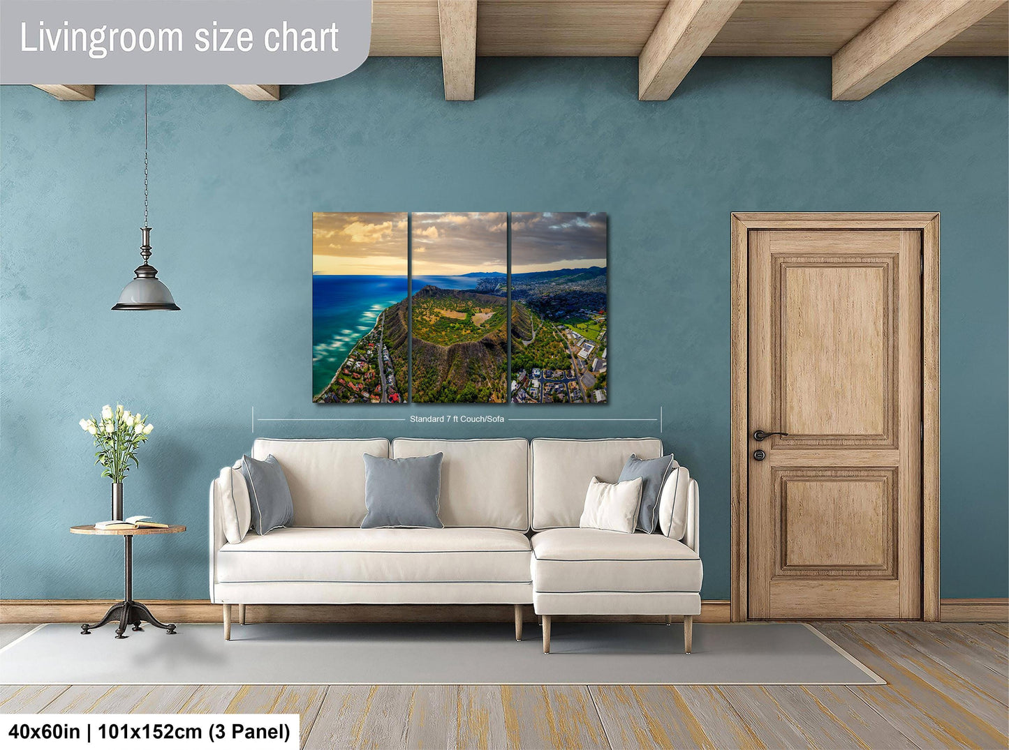 The image is a living room with a large, colorful, and detailed landscape painting hanging on the wall. The painting depicts a scenic coastal area with mountains, water, and greenery. The room has a white sofa, a wooden door, and a small table with a vase of flowers. The painting is approximately 40 inches wide and 101.5 inches tall, with a 3-panel layout.