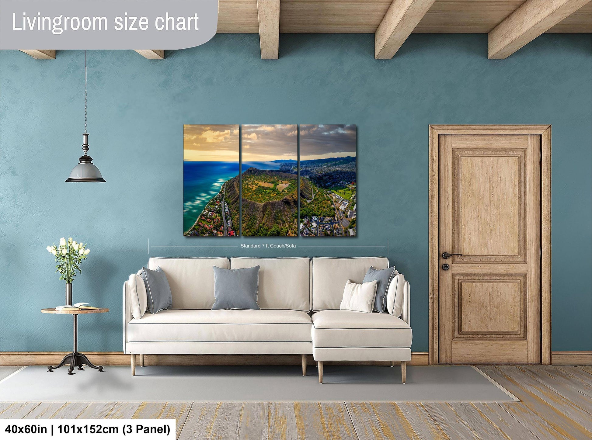 The image is a living room with a large, colorful, and detailed landscape painting hanging on the wall. The painting depicts a scenic coastal area with mountains, water, and greenery. The room has a white sofa, a wooden door, and a small table with a vase of flowers. The painting is approximately 40 inches wide and 101.5 inches tall, with a 3-panel layout.